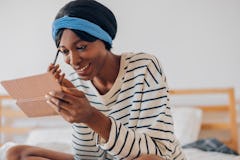 A portrait of a smiling black woman putting on makeup while sitting on the bed before work