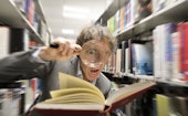 Man with magnifying glass in library looking astonished at facts in a bookAcademic