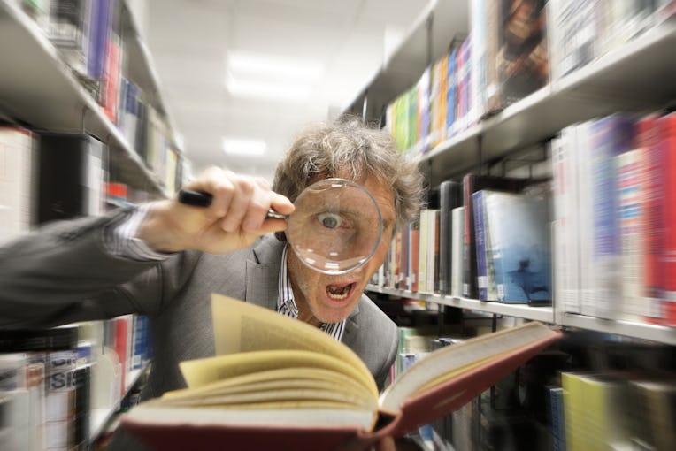 Man with magnifying glass in library looking astonished at facts in a bookAcademic