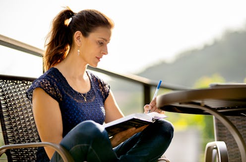 Woman writing in her journal while sitting cross-legged in a chair outside on her patio on a sunny d...