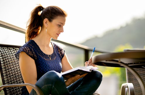 Woman writing in her journal while sitting cross-legged in a chair outside on her patio on a sunny d...