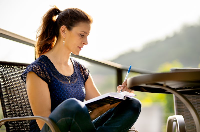 Woman writing in her journal while sitting cross-legged in a chair outside on her patio on a sunny d...