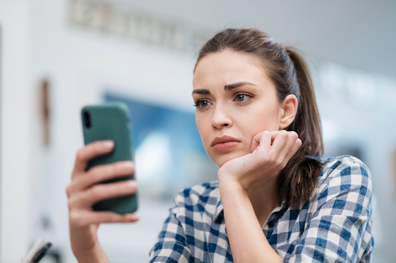 Close up of a depressed Caucasian young woman sitting at home, reading some bad news on her smart ph...