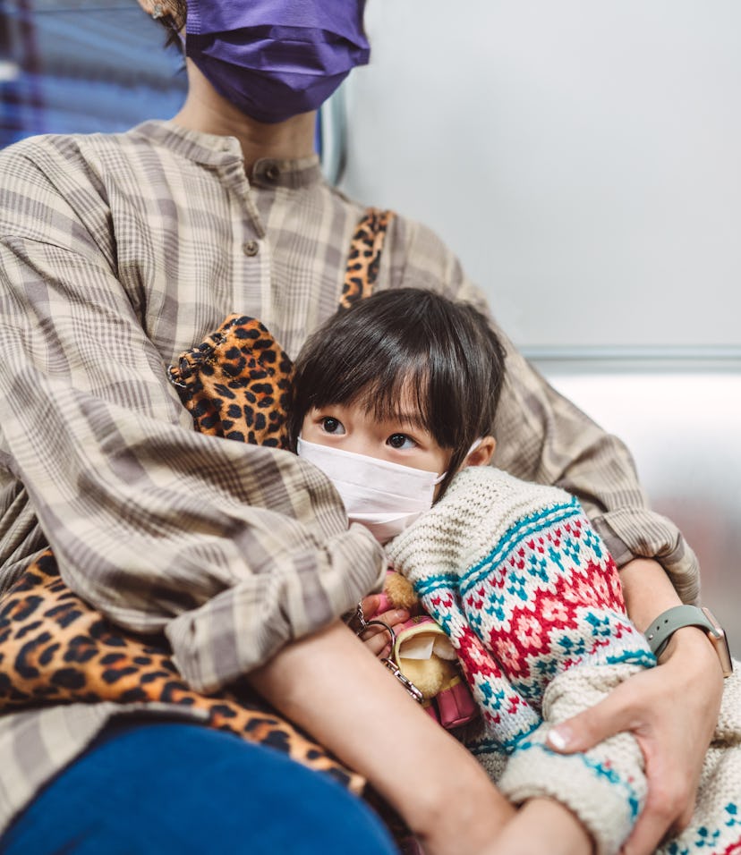 Lovely little girl in protective face mask resting on her mom’s arm while riding on train.