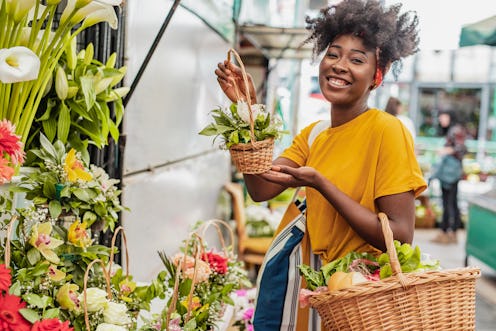 A happy smiling African-American woman is holding a basket full of groceries and buying flowers