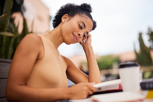 Smiling young female university student sitting outside at a table on campus drinking coffee and rea...