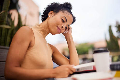 Smiling young female university student sitting outside at a table on campus drinking coffee and rea...