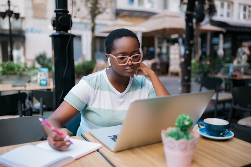 Young African American female freelancer sitting in a coffee shop, using a laptop for work while wri...