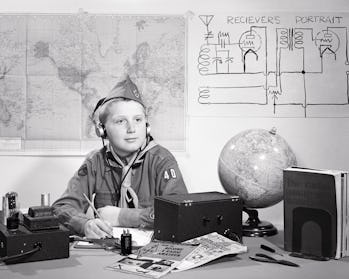 1950s boy scout in uniform a amateur ham radio operator sitting at desk wearing earphones listening ...