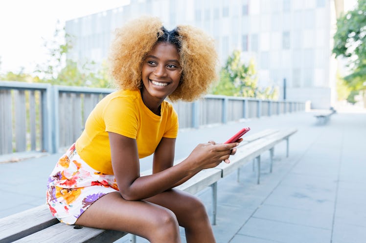 Trendy young afro-american woman using smart phone while sitting on bench in city street - Happy mix...