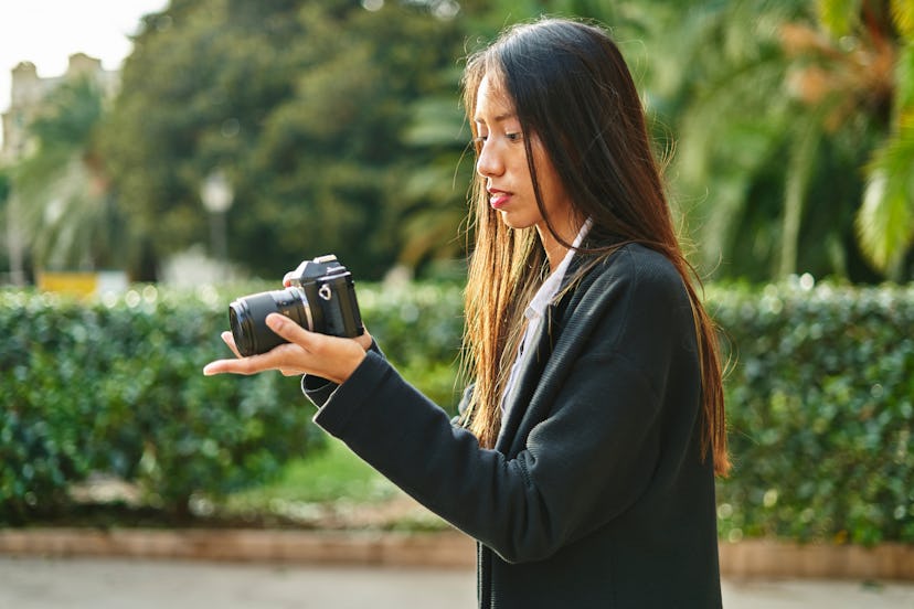 Focused Hispanic female in stylish outfit taking photo on modern photo camera in hands standing on s...