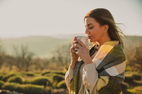 Portrait of beautiful young woman standing outside, in warm autumnal sunshine, wrapped in a cozy sca...
