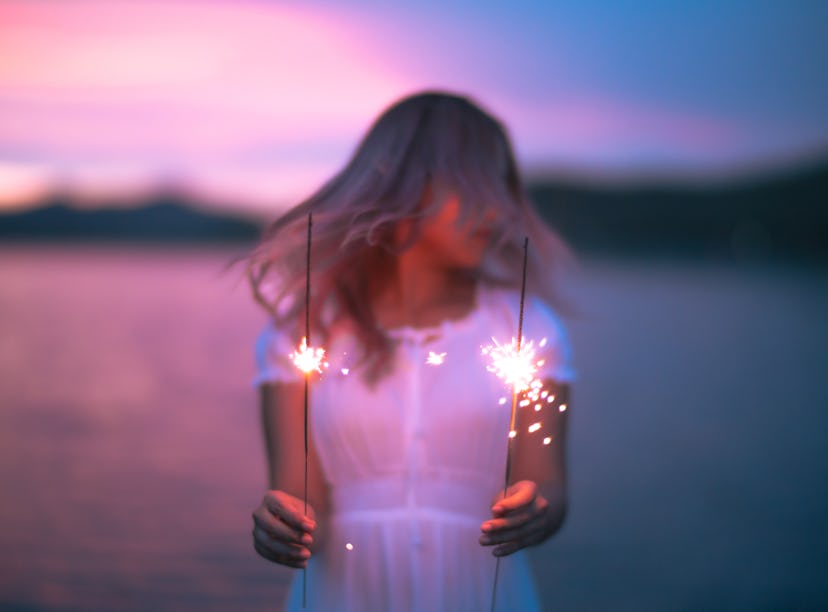 Young woman holding two sparklers by the ocean at sunset, thinking about how her zodiac sign is the ...