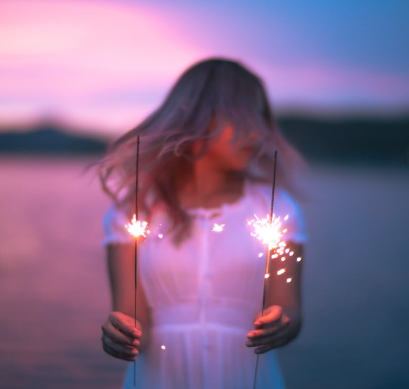 Young woman holding two sparklers by the ocean at sunset, thinking about how her zodiac sign is the ...
