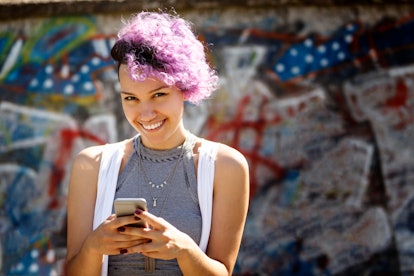 A smiling woman in front of a graffiti wall types in some manifestation quotes as captions on Instag...