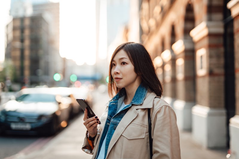 Young Asian woman using mobile app on smartphone to order taxi pick up service in the city. She is l...