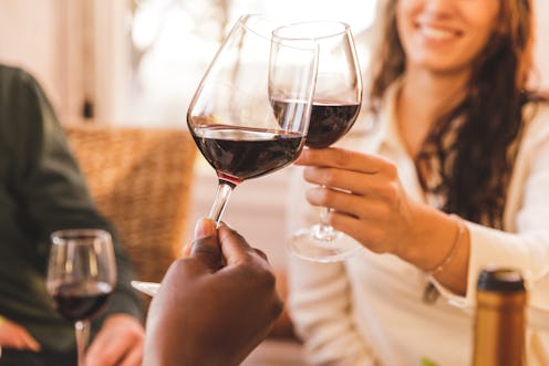 Close up shot of smiling young woman making a celebratory toast with red wine, with a friend sitting...