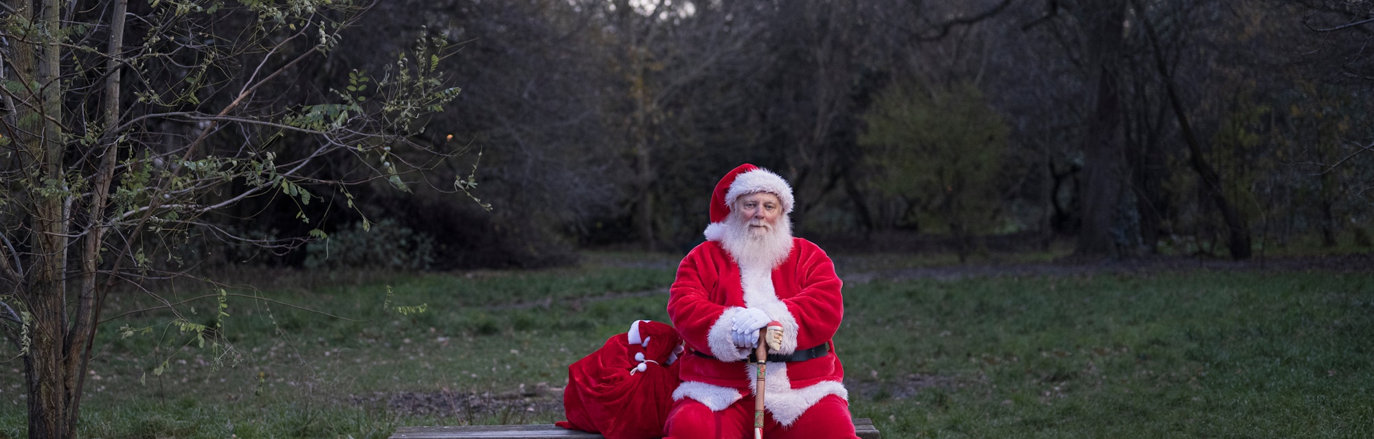 Full length front view of bearded senior man sitting on park bench with sack full of gifts and looki...