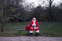 Full length front view of bearded senior man sitting on park bench with sack full of gifts and looki...