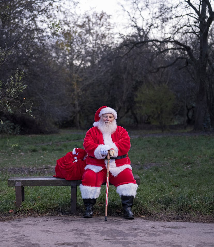 Full length front view of bearded senior man sitting on park bench with sack full of gifts and looki...