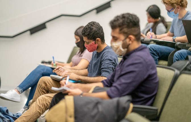 College students sitting in a lecture hall on campus, all are wearing masks to protect from the tran...