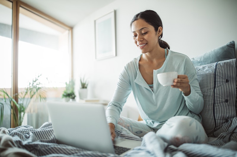 Woman in bed working something on laptop and drinking tea