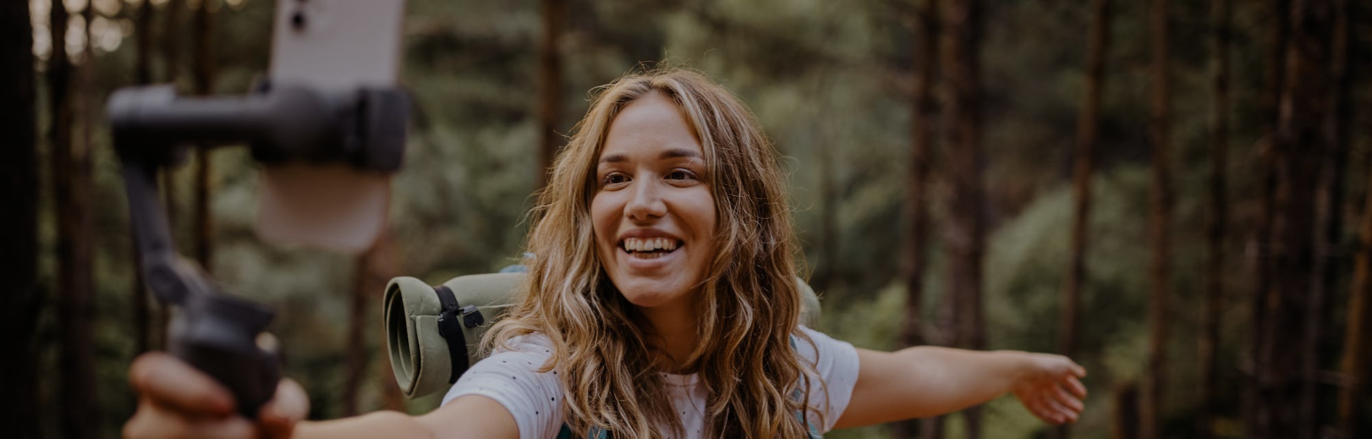 Cheerful young female vlogger with backpack during hiking adventure recording vlog and going live fo...