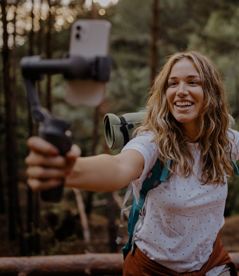 Cheerful young female vlogger with backpack during hiking adventure recording vlog and going live fo...
