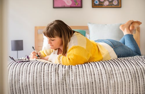 A woman in a yellow sweater journals in bed. Here's your daily horoscope for December 20, 2021.