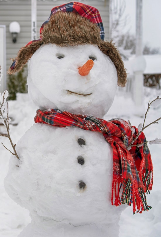 Cute smiling snowman with red scarf and hat in front of a snowy porch on a winter day