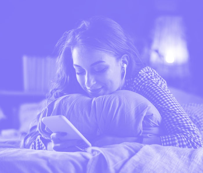 Portrait of smiling teenage girl lying on front on her cozy bed, embracing a pillow, listening to mu...
