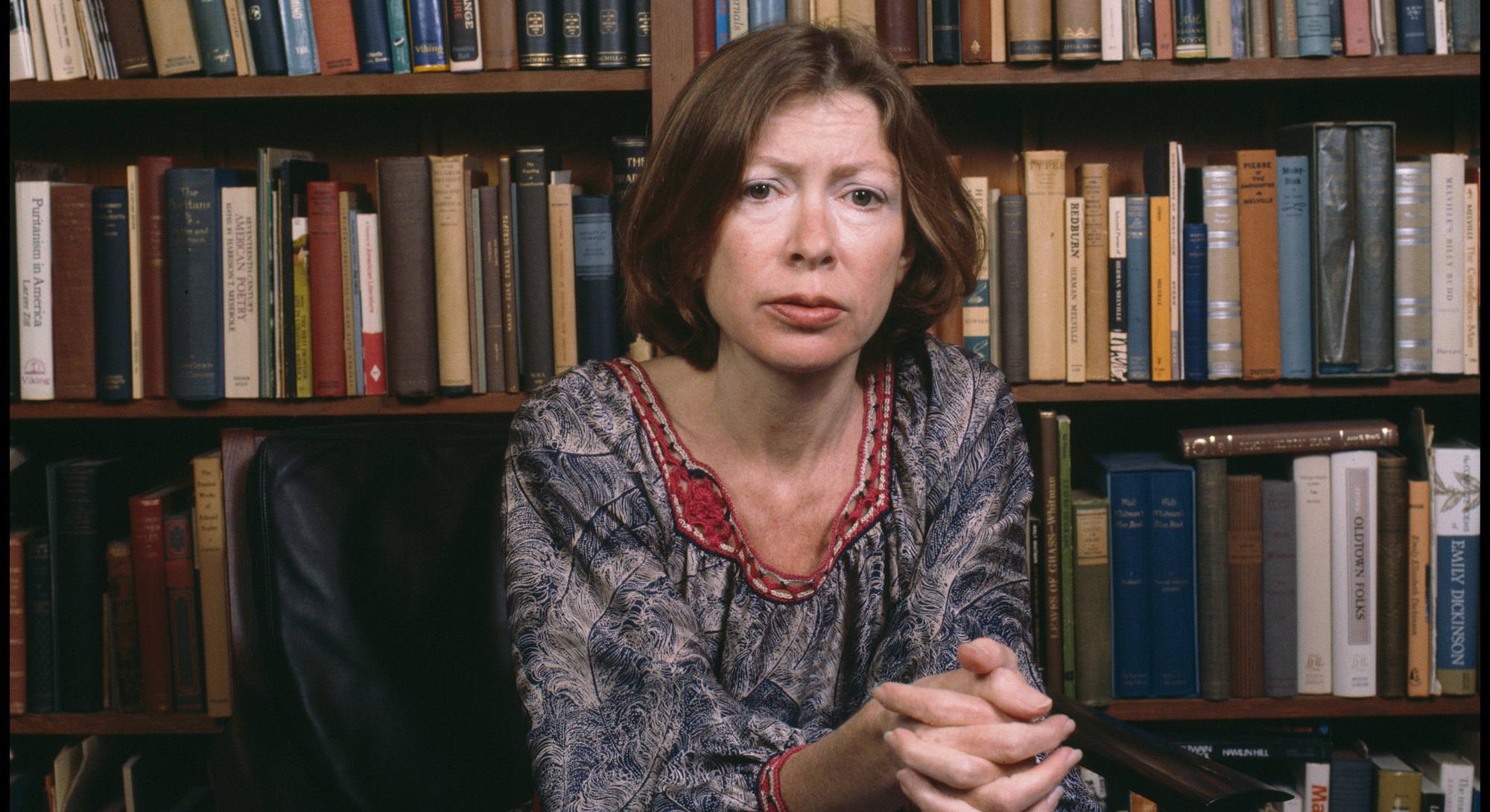 Portrait of American author Joan Didion as she sits in a chair in front of a bookshelf, Berkeley, Ca...