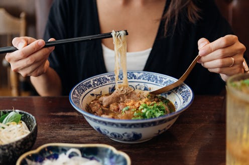 Close-up shot of young Asian woman eating Chinese noodle soup with chopsticks and spoon in restauran...