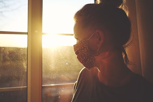 Pretty woman in quarantine with mask, looks out the window