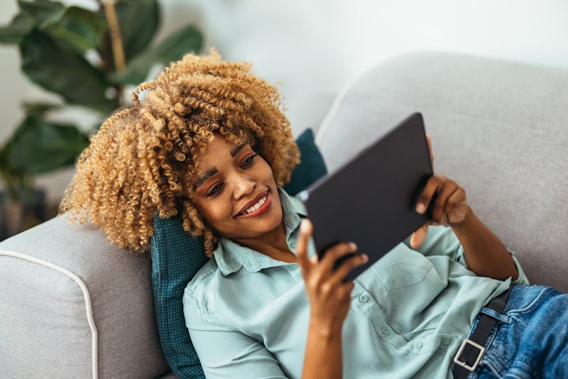 Smiling Afro-American woman is lying on the sofa and watching something on a digital tablet. She is ...