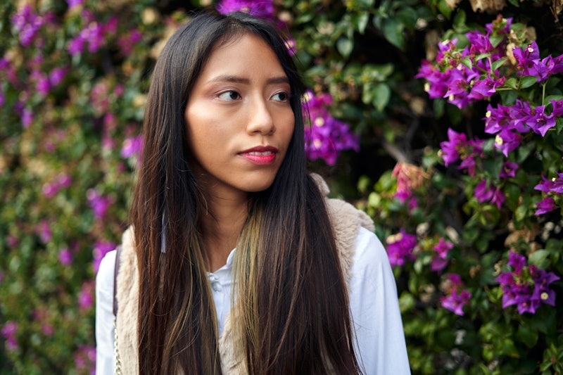 Charming Hispanic female with long hair looking away while standing near blossoming tree with green ...