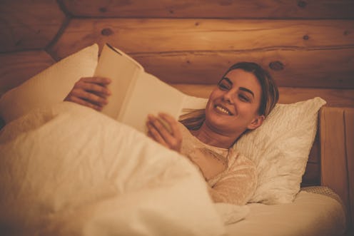 Low angle view of a happy woman lying on a bed and reading before going to sleep.
