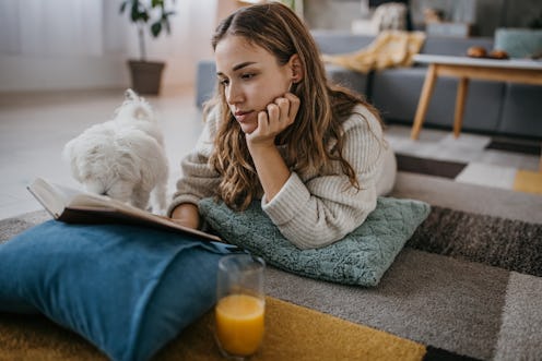 Young woman lying on the floor and reading a book, her Maltese pet dog is with her at home.