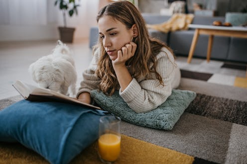 Young woman lying on the floor and reading a book, her Maltese pet dog is with her at home.