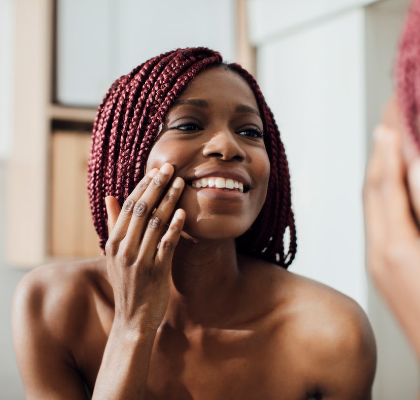 Beautiful smiling African woman standing in front of a mirror and looking at her face wrinkles.