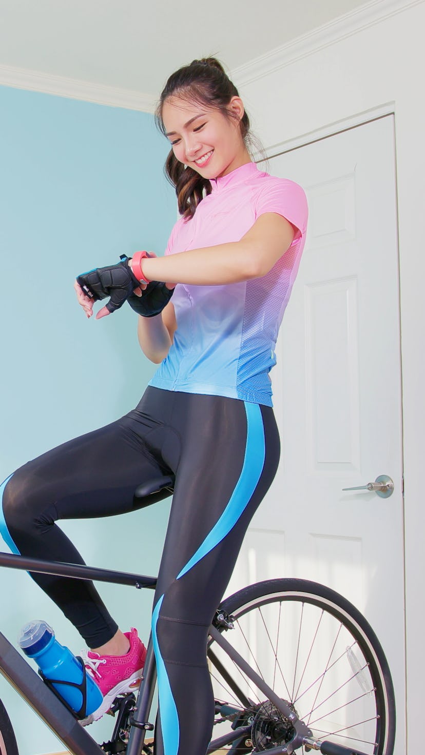 A woman rides on a home bike and checks her speed. Fitness pros recommend strength training for cycl...