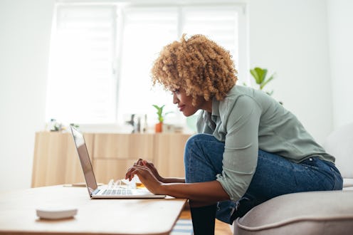 Side view of a smiling Afro-American businesswoman who is sitting on a sofa and typing on a laptop o...