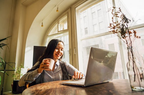 A middle-aged asian woman in blue denim shirt sitting at the table in front of a laptop, drinking a ...
