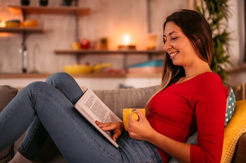 Young beautiful woman lying on sofa at home, drinking coffee and reading book in the evening.
