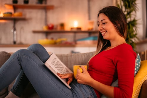 Young beautiful woman lying on sofa at home, drinking coffee and reading book in the evening.