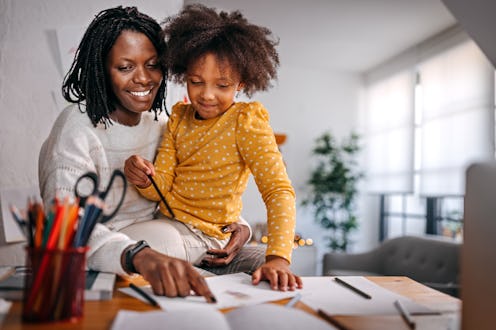 Cheerful and happy mother with curly hair helping cute little daughter in drawing on paper with colo...