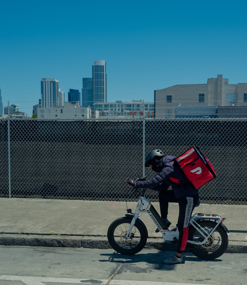 Doordash driver on electric bicycle wearing a cooler backpack and consulting phone with skyline of S...