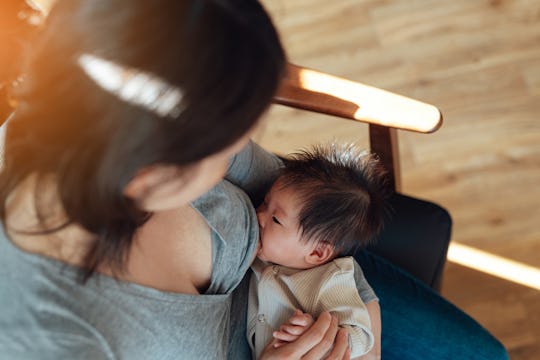 Over the shoulder view of young Asian mom breastfeeding newborn baby while sitting on armchair at ho...