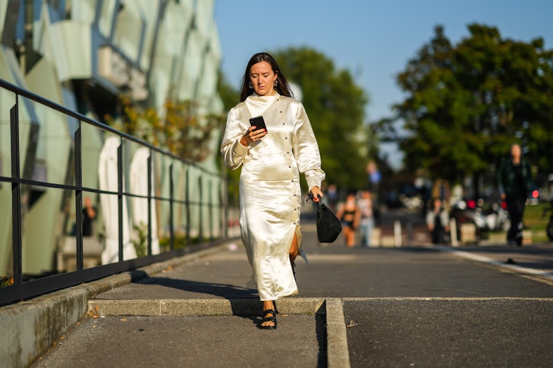 A woman in a shiny white dress shares someone else's Instagram story while walking down the street. 