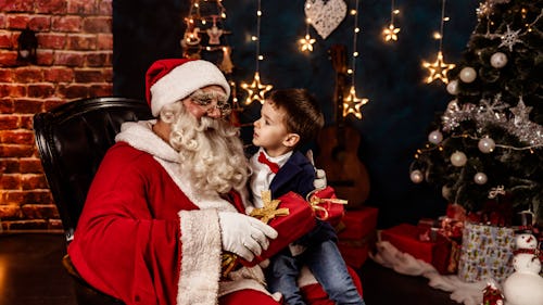 Photo of  little boy sitting in Santa's lap at home and talking to him while holding Christmas prese...